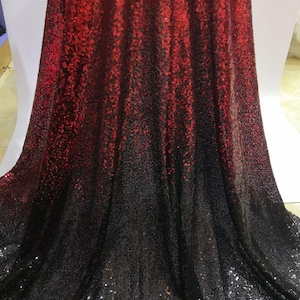 Peut inclure: Un tissu avec un effet dégradé, passant du rouge en haut au noir en bas. Le matériau est recouvert de petites paillettes réfléchissantes, créant une surface chatoyante et texturée. Le tissu est drapé, suggérant qu'il pourrait être utilisé pour des vêtements ou à des fins décoratives.