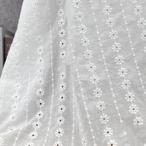 Pode incluir: Close-up de tecido branco com bordado inglês e padrão floral. O tecido apresenta filas de pequenas flores bordadas e linhas verticais de pequenos pontos. O tecido de bordado inglês é provavelmente usado para roupas ou decoração de casa.