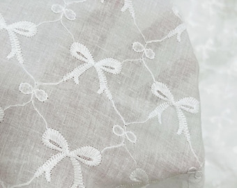 May include: Close-up of white fabric with embroidered bow and loop patterns. The delicate design features repeating bows connected by thin lines, creating a lace-like effect. The fabric is slightly translucent, suggesting a lightweight material.