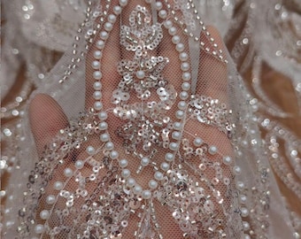 Puede incluir: Primer plano de tela de encaje marfil adornada con lentejuelas y perlas de imitación. La delicada red está adornada con intrincados patrones de lentejuelas brillantes y pequeñas perlas redondas, creando una textura lujosa y elegante. Ideal para novias o ropa formal.