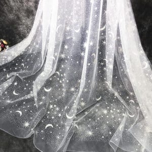 May include: White sheer fabric featuring a celestial design of stars and crescent moons. The fabric is draped, creating soft folds and a flowing appearance. The design is a repeating pattern of various sized stars and moons.