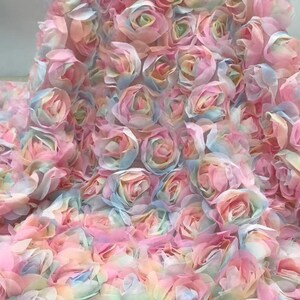 May include: A close-up view of a fabric covered in pastel-colored, three-dimensional roses. The roses are primarily pink, with hints of blue, yellow, and green. The fabric appears to be delicate and suitable for decorative purposes.