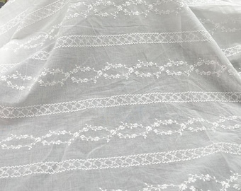 May include: White fabric with embroidered floral and geometric patterns. The fabric is folded, showing the texture and design details. The embroidery is delicate and adds visual interest to the textile.