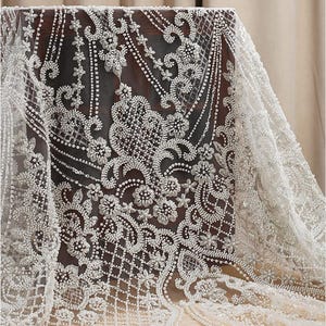 May include: Close-up of white beaded lace fabric. The intricate design features floral and geometric patterns, with small beads creating texture and dimension. The lace is draped over a dark surface, showcasing the delicate details.
