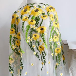 Peut inclure: Tissu blanc transparent orné de broderies de tournesols jaunes, de feuilles vertes et de petites fleurs blanches. Le motif floral est disposé en cascade, créant un effet délicat et élégant. Idéal pour les voiles de mariée ou les projets décoratifs.