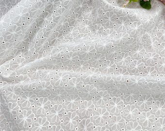 May include: White eyelet fabric with a floral pattern. The fabric is a light, airy material with embroidered flower designs and small cut-out holes. The fabric is draped, creating soft folds and shadows.