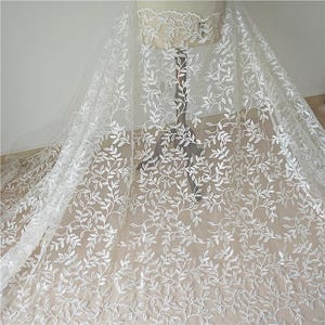 Peut inclure: Tissu en dentelle blanche avec un motif de feuilles délicat. Le tissu est drapé sur un mannequin, mettant en valeur le design floral complexe. La dentelle est semi-transparente, avec un motif de feuilles en relief.