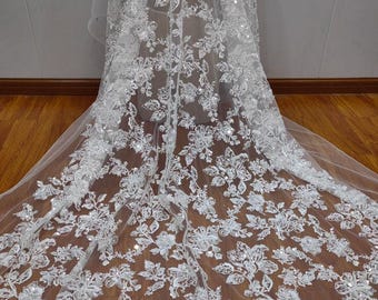 以下が含まれることがあります： 白のフローラルレース生地のクローズアップ。複雑な刺繍のディテールとキラキラ光るスパンコールが特徴です。繊細なデザインは、花と葉の繰り返し模様で、質感とエレガントな外観を作り出しています。ブライダルウェアや特別な機会の衣服に最適です。