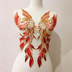以下が含まれることがあります： 鮮やかな赤、オレンジ、金の糸で刺繍された左右対称のフェニックスのワッペン。鳥のモチーフには複雑な翼の羽と、青いアクセントが入った長く流れるような尾羽が詳細に描かれており、布地に縫い付けたり貼り付けたりして使用するデザインです。