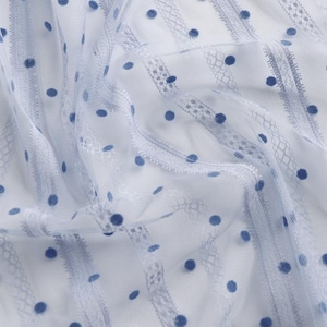 May include: Close-up of a white fabric with a delicate, sheer texture. The fabric features a pattern of blue polka dots and vertical stripes with intricate lace detailing, creating a soft and airy aesthetic. Ideal for apparel or home decor.