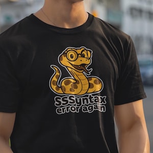 Puede incluir: Camiseta negra con una serpiente de dibujos animados con gafas y el texto "SSSyntax error again". La serpiente es amarilla y marrón. La camiseta se combina con pantalones blancos. La foto está tomada al aire libre.