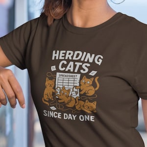 以下が含まれることがあります： 「HERDING CATS SPREADSHEET SINCE DAY ONE」の文字と猫のグラフィックがプリントされた茶色のTシャツ。フープイヤリングをした人が着ています。