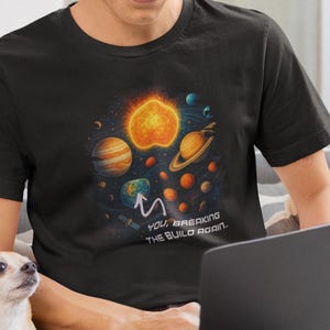 Könnte beinhalten: Schwarzes T-Shirt mit einer farbenfrohen Illustration von Planeten, Sternen und einer Sonne. Der Text "YOU ARE BREAKING THE BUILD AGAIN" ist darunter gedruckt. Eine Person sitzt mit einem Laptop und einem kleinen Hund.