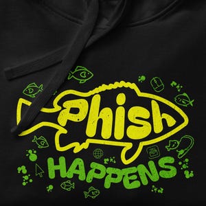 May include: Black hooded sweatshirt with a drawstring. The front features a yellow and green graphic of a fish with the words "Phish Happens" in a playful font. Small fish and other icons surround the text.