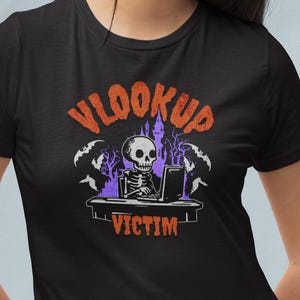 May include: Black t-shirt featuring a Halloween-themed graphic. The design includes a skeleton at a computer, bats, and the words "VLOOKUP VICTIM" in orange and purple. The shirt is made of a soft material.