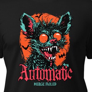 Könnte beinhalten: Schwarzes T-Shirt mit einem Monster-Grafikdesign. Das Design zeigt ein Monster mit roten Augen, scharfen Zähnen und türkisfarbenem Fell vor einem orangefarbenen Hintergrund. Der Text "Automatic" ist pink, darunter steht "Merge Failed".