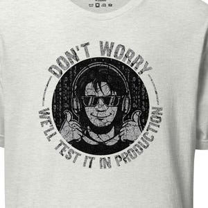 Puede incluir: Camiseta gris claro con un gráfico desgastado. El gráfico presenta a una persona con auriculares y gafas de sol, haciendo una señal de pulgar hacia arriba, rodeada por el texto "DON'T WORRY, WE'LL TEST IT IN PRODUCTION."