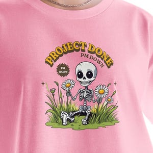 Puede incluir: Camiseta rosa con un gráfico de un esqueleto de dibujos animados sosteniendo margaritas, con el texto "PROJECT DONE PM DOWN" en una fuente retro. El esqueleto está en un campo de hierba y flores. La camiseta se está usando.