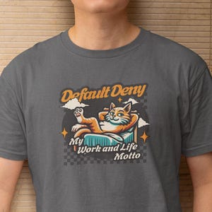 May include: A grey t-shirt featuring the words "Default Deny" and a cartoon cat illustration. The cat is relaxing on a pillow with the words "My Work and Life Motto" underneath. The t-shirt is worn by a person.