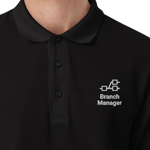 Könnte beinhalten: Schwarzes Poloshirt mit einem weißen, gestickten Logo und dem Schriftzug "Branch Manager". Das Shirt hat einen klassischen Kragen und eine Knopfleiste mit drei Knöpfen. Die kurzen Ärmel haben ein kleines weißes Logo.