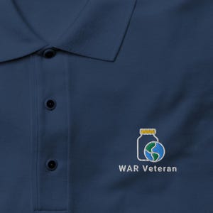 May include: Navy blue polo shirt with a collar and three-button placket. The shirt features an embroidered design of a jar and globe with the text "WAR Veteran" in white. The shirt is from Port Authority.