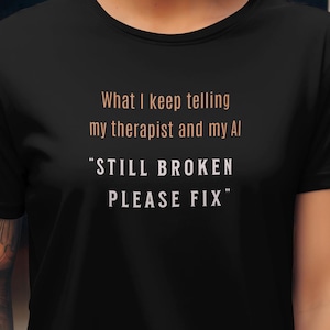 Peut inclure: T-shirt noir avec le texte "What I keep telling my therapist and my AI" et "STILL BROKEN PLEASE FIX" en blanc et orange. Le t-shirt est de style col rond.