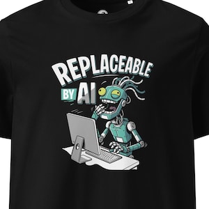 Può includere: Una t-shirt nera a maniche corte con la grafica di un robot color verde acqua e grigio seduto davanti a un computer. Il robot ha un'espressione ridente. Sopra il robot, il testo "REPLACEABLE BY AI" è scritto in lettere maiuscole bianche e verde acqua.