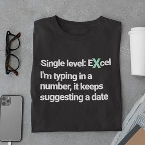 Pode incluir: Camiseta preta com o texto "Single level: Excel, I'm typing in a number, it keeps suggesting a date." A imagem também inclui um smartphone, óculos, um copo de café e um jornal.
