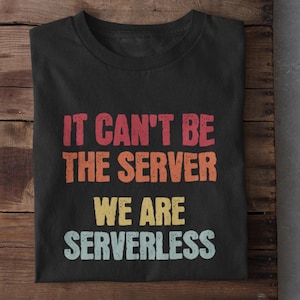 May include: Black t-shirt with the text "IT CAN'T BE THE SERVER WE ARE SERVERLESS" in red, orange, yellow, and blue. The shirt is folded on a wooden surface, with white trainers visible at the bottom.