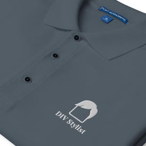 May include: A slate grey polo shirt with a collar and three buttons. The shirt features a white embroidered logo and the text "DIY Stylist". The tag reads "Port Authority".