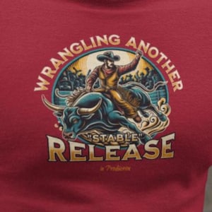 May include: A red t-shirt with a rodeo-themed design. The graphic shows a cowboy on a bull, with the text "WRANGLING ANOTHER STABLE RELEASE". The shirt is made of a soft material and has short sleeves. The design is in a circular shape.