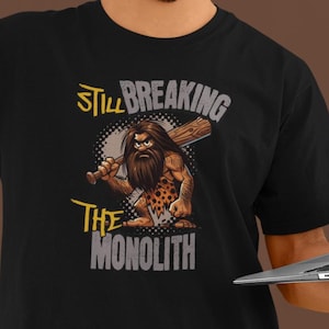 May include: Black t-shirt featuring a cartoon caveman graphic with the text "Still Breaking The Monolith". The person in the image is holding a laptop.