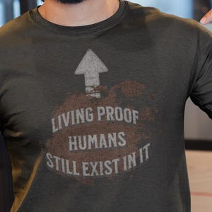 Pode incluir: Uma camiseta verde escuro com o texto "LIVING PROOF HUMANS STILL EXIST IN IT" e um gráfico de seta. A pessoa na imagem está usando um boné de beisebol azul e jeans azuis.