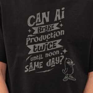 Op de afbeelding: Zwart T-shirt met de tekst "CAN AI BREAK PRODUCTION TWICE until noon SAME DAY?" in een vervaagde grijze letter. Het shirt heeft een kleine cartoon eend. Het model draagt een zwarte pet en zonnebril.