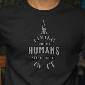 Könnte beinhalten: Schwarzes T-Shirt mit dem Text "Living Proof Humans Still Exists In It" in weißer, stilisierter Schrift. Die Person im Bild trägt eine blaue Jeans und hält eine Zeitung.