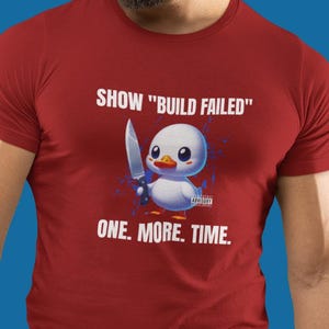 Puede incluir: Una camiseta roja con el texto "SHOW 'BUILD FAILED'" y "ONE. MORE. TIME." presenta un pato de dibujos animados sosteniendo un cuchillo. La camiseta la lleva una persona. El fondo es azul.