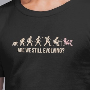 Può includere: T-shirt nera con una grafica umoristica che mostra l'evoluzione umana, che culmina con una persona che usa un laptop. Il testo "ARE WE STILL EVOLVING?" è sotto. Un design divertente e stimolante.
