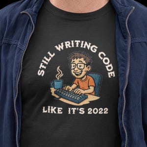 以下が含まれることがあります： 「Still Writing Code Like It's 2022」の文字と、プログラミングをしている人の漫画のグラフィックが描かれた黒いTシャツ。メガネをかけ、コーヒーカップを持ってキーボードの前に座っている。オレンジ色のシャツを着ている。