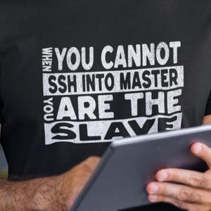 Può includere: T-shirt nero con la scritta bianca "WHEN YOU CANNOT SSH INTO MASTER ARE THE SLAVE". La persona tiene in mano un tablet. La maglietta è un capo casual.