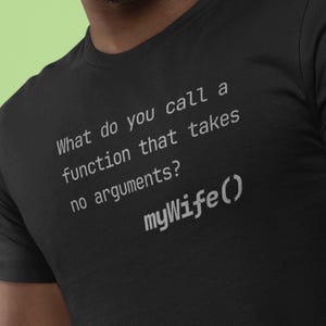 Puede incluir: Camiseta negra con texto blanco. El texto dice: "¿Cómo llamas a una función que no toma argumentos? myWife()". La camiseta es de cuello redondo.
