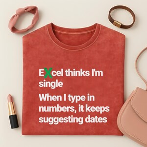 Op de afbeelding: Een roestkleurig t-shirt met de tekst "Excel denkt dat ik single ben. Als ik getallen invoer, blijft het data voorstellen." Een roze haarband, lippenstift, een bruine armband en een roze handtas staan ook op de afbeelding.