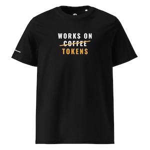 May include: A black short-sleeve t-shirt featuring the white text WORKS ON, the word COFFEE in white with a horizontal orange line through it, and the word TOKENS in orange below. The shirt has a crew neckline and a small logo on the sleeve.