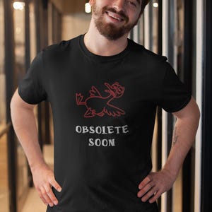 Op de afbeelding: Zwart t-shirt met een rode cartoon eend ontwerp en de witte tekst "OBSOLETE SOON". Het t-shirt heeft een ronde hals en is gemaakt van een zachte stof.