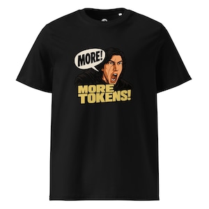 May include: A black short-sleeved t-shirt featuring a graphic illustration of a man with dark hair and a scar on his face, shouting with his mouth open. Above him is a white speech bubble with the black text MORE! and the yellow text MORE TOKENS!