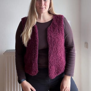 Könnte beinhalten: Eine weinrote Weste mit strukturiertem, flauschigem Aussehen. Die Weste wird über einem langärmeligen braunen Oberteil und einer schwarzen Hose getragen. Die Weste hat einen V-Ausschnitt und scheint aus einem weichen, warmen Material zu sein. Der Gesamtstil ist lässig und bequem.