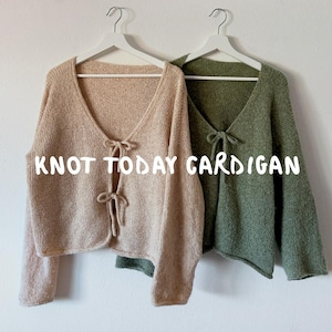Może przedstawiać: Dwa kardigany z otwartym przodem wiszące na białych wieszakach. Jeden jest w kolorze jasnobeżowym, a drugi oliwkowozielonym. Oba mają wiązanie z przodu. Tekst "KNOT TODAY CARDIGAN" jest wyświetlany na biało.
