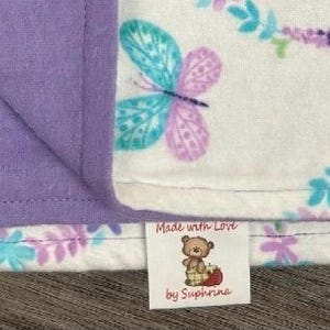 May include: A white and purple floral print blanket with a lavender backing. The blanket has a tag that reads "Made with Love by Suphrina".