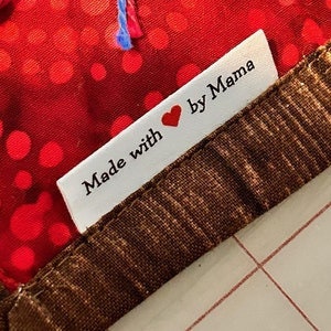 May include: A close-up of a white tag with the text "Made with ❤️ by Mama" in black font. The tag is sewn onto a brown patterned fabric and a red fabric with red polka dots.