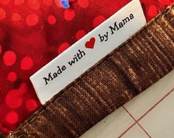 May include: A close-up of a white tag with the text "Made with ❤️ by Mama" in black font. The tag is sewn onto a brown patterned fabric and a red fabric with red polka dots.