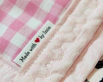 May include: A textile item featuring a pink and white gingham pattern on one side and a solid light pink fabric with a wavy quilted stitch pattern on the other. A white fabric label is sewn onto the edge with the text Made with heart by Izzie.